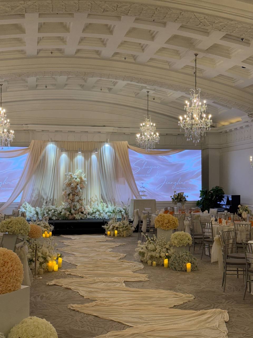 An ivory fabric aisle winding through white florals beneath ballroom chandeliers