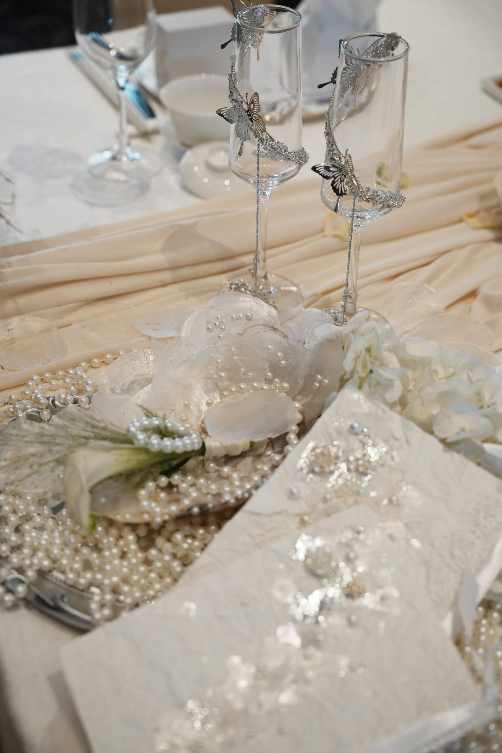 Butterfly-adorned champagne flutes, pearl details and white flowers on ivory fabric